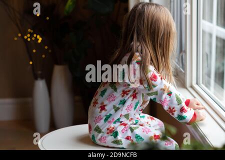 young girl looks out window while wearing Christmas pyjamas Stock Photo