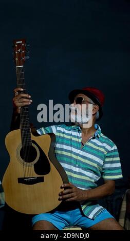 Bearded Asian senior man wearing a hat and sunglasses posing at the camera with a guitar Stock Photo