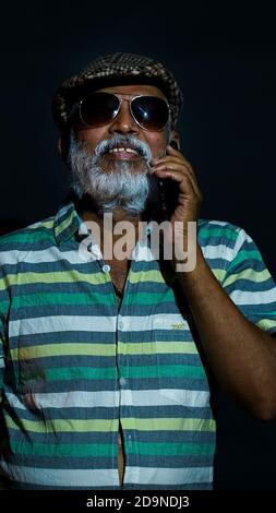 Happy bearded Asian senior man wearing a hat and sunglasses talking on the phone Stock Photo