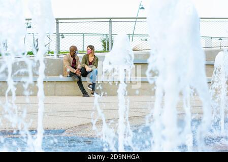 multiethnical couple wearing protective face mask interacting with each other sitting in a park bench with a beautiful fountain splash in autumn Stock Photo
