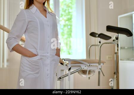 Closeup of a doctor in a white uniform, with folded hands in front of him. Stands in front of a gynecological chair. Stock Photo