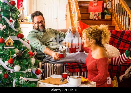 Christmas time at home with happy adult caucasian couple having breakfast on the couch near the christmas tree - concept of family and love life together in traditional leisure activity indoor