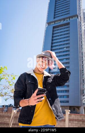 Cheerful alternative young man teenager use cellular phone to write - skyline building in the city background - concept of real young teeanger lifestyle and connection device - violet diversity hair and clothes Stock Photo