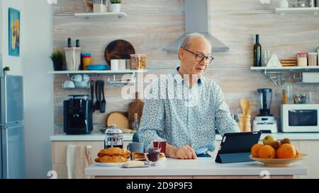 Happy senior man during breakfast in kitchen having a video conference with family. Elderly person using internet online chat technology video webcam making a video call connection camera communication conference call Stock Photo