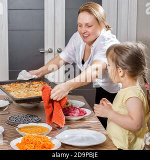 Mom puts a tray of hot homemade pizza on the table. little daughter looks with appetite. dinner with family Stock Photo