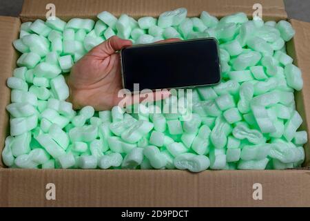 A hand holding a smartphone emerges from a cardboard box full of polystyrene chips Stock Photo