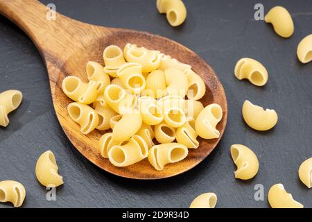 Uncooked elbows macaroni pasta in wooden spoon on dark table. Stock Photo