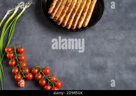Grilled, whole, delicious sausages are on the frying pan, which stands on a gray background. Nearby are two branches of fresh cherry tomatoes and onio Stock Photo
