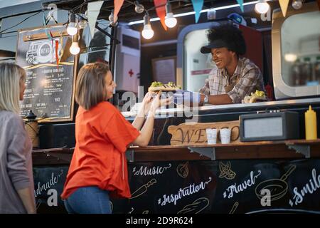 two caucasian females standing in line for fast food service. Young afro-american employee giving from hand to hand sandwiches, wearing gloves, smilin Stock Photo