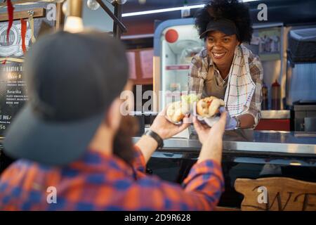 close up of sandwiches with green salad and mustard. caucasin male receiving from a smiling afro-american employee in fast food service Stock Photo