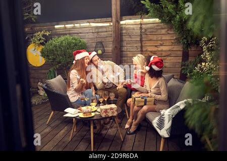 group of young adult friends celebrating new year outdoor, sitting in the backyard with sparkling sticks in their hands wearing santa hats Stock Photo