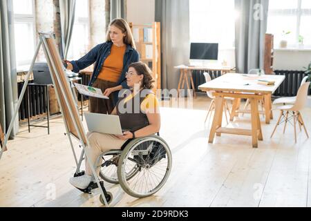 Young businesswoman in casualwear helping disable colleague with presentation Stock Photo