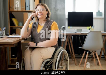 Happy young female agent or secretary in wheelchair consulting on mobile phone Stock Photo