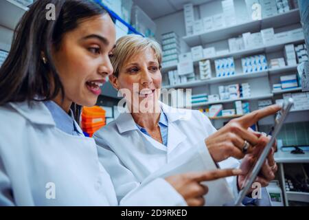 Elderly female pharmacist guiding new intern scrolling through names of medication on digital tablet  Stock Photo