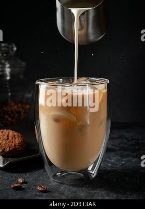 Pouring cream into the glass of iced coffee on black background. Stock Photo