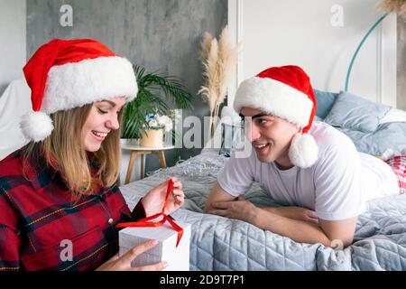 Christmas, holidays, winter and people concept. Happy couple with gift present box at home wearing santa hats. Young smiling man lying on the bed give Stock Photo