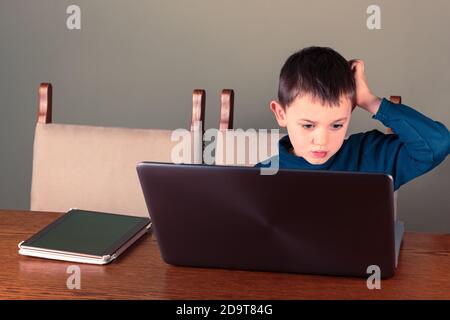 Eight year old boy using laptop and tablet computers scratches his head. Homeschooling or digital technology theme Stock Photo