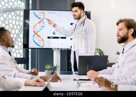 Genetic technology concept, gene engineering. Young handsome concentrated Indian male doctor showing his multiethnic colleagues the dna molecule on Stock Photo