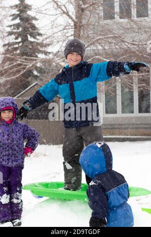 Child standing on sled in snow smiling and looking at camera while siblings play near him Stock Photo