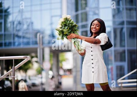 Beautiful african american girl holding bouquet of white roses flowers on dating in the city. Black businesswoman with bunch of flowers. Stock Photo