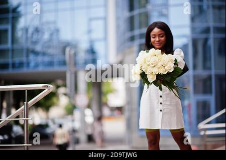 Beautiful african american girl holding bouquet of white roses flowers on dating in the city. Black businesswoman with bunch of flowers. Stock Photo