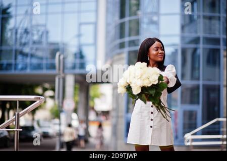 Beautiful african american girl holding bouquet of white roses flowers on dating in the city. Black businesswoman with bunch of flowers. Stock Photo