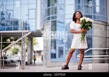 Beautiful african american girl holding bouquet of white roses flowers on dating in the city. Black businesswoman with bunch of flowers. Stock Photo