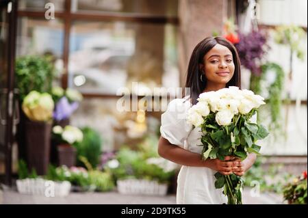 Beautiful african american girl holding bouquet of white roses flowers on dating in the city. Black businesswoman with bunch of flowers. Stock Photo