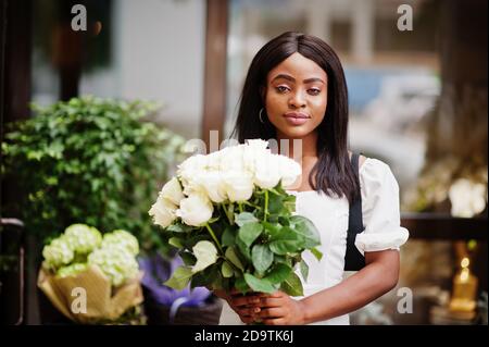 Beautiful african american girl holding bouquet of white roses flowers on dating in the city. Black businesswoman with bunch of flowers. Stock Photo