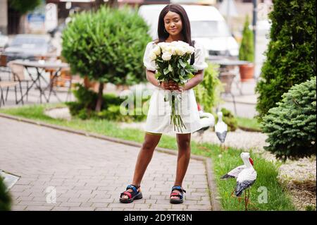 Beautiful african american girl holding bouquet of white roses flowers on dating in the city. Black businesswoman with bunch of flowers. Stock Photo