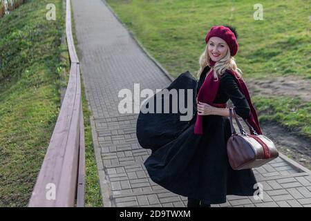 A happy woman walks up the stairs in burgundy palette and biret, an adult with a beautiful smile in black clothes, in the fall against the background Stock Photo