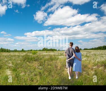 Back view of friendly girl and guy holding hands walking in the forest with sky with bright white clouds. Happy young couple walking together through a wheat field. Back view people collection. Stock Photo