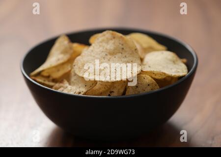 Organic potato chips with black pepper in black ceramic bowl on walnut wood background , shallow focus Stock Photo