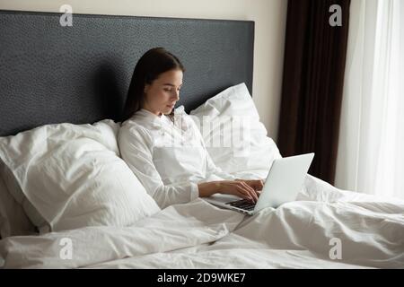 Concentrated millennial woman working online at laptop in bed Stock Photo