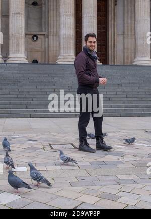 A man standing at St Paul’s Cathedral west front smiling. Pigeons on the ground and steps and pillars in background. Stock Photo