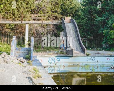 An old abandoned outdoor pool with a slide and bridge and green swampy water is located in the middle of the park Stock Photo