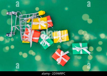 A fallen small cart with scattered gifts. Red, green and yellow gifts in a shopping trolley on green background with bokeh. Christmas and Christmas gift concept. Top view Stock Photo
