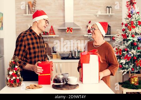 Happy grandparents celebrating christmas exchanging presents with red bow. Senior man and woman wearing santa hat during christmas giving elderly woman gift box with christmastree in the background. Stock Photo