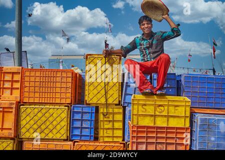A young Indian man sitting on colorful plastic crates in the Old Port of Mangalore, Karnataka, India, lifts his hat in a cheerful greeting Stock Photo
