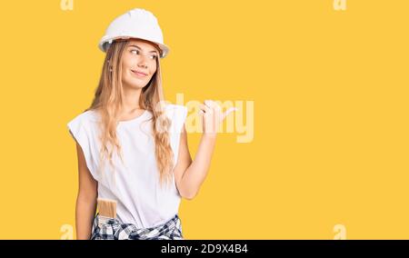 Beautiful caucasian woman with blonde hair wearing hardhat and painter clothes smiling with happy face looking and pointing to the side with thumb up. Stock Photo