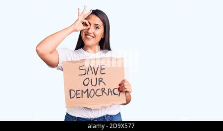 Young beautiful woman holding save our democracy cardboard banner smiling happy doing ok sign with hand on eye looking through fingers Stock Photo