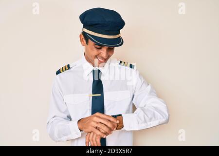 Young hispanic man wearing airplane pilot uniform checking the time on wrist watch, relaxed and confident Stock Photo