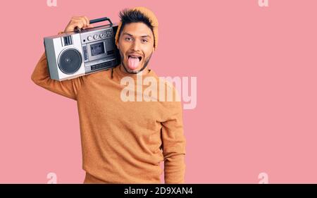 Handsome latin american young man holding boombox, listening to music sticking tongue out happy with funny expression. emotion concept. Stock Photo
