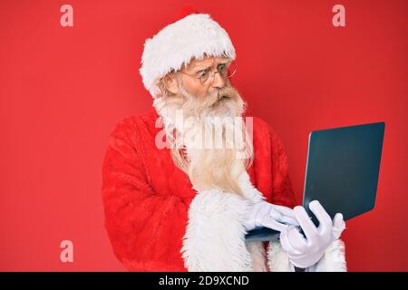 Old senior man with grey hair and long beard wearing santa claus costume using laptop in shock face, looking skeptical and sarcastic, surprised with o Stock Photo