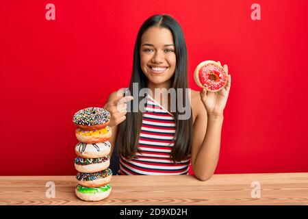Beautiful hispanic woman holding tasty colorful doughnuts smiling happy pointing with hand and finger Stock Photo