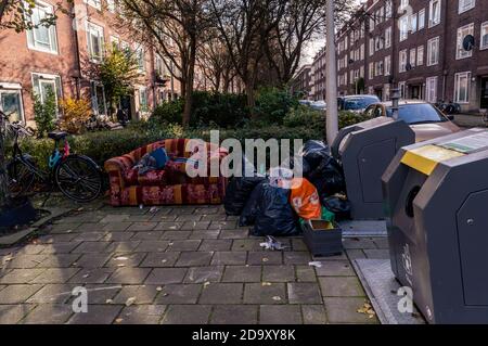 AMSTERDAM, THE NETHERLANDS OCTOBER 21, 2020: A discarded colorful couch and a heap of garbage piled up against a public dumpster in a street Stock Photo