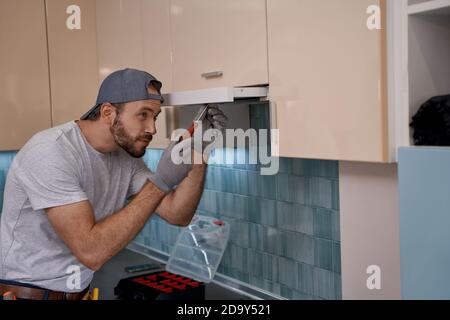 Young caucasian repairman installing kitchen hood using screwdriver Stock Photo