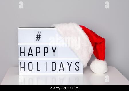 Lightbox with happy holidays hashtag text and red Santa hat on white table near the wall Stock Photo