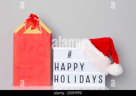 Lightbox with happy holidays hashtag text wearing Santa hat and paper shopping bag filled with Christmas gifts on white table Stock Photo