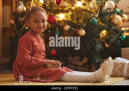Warm toned portrait of smiling African-American girl opening Christmas presents while sitting by tree at home, copy space Stock Photo